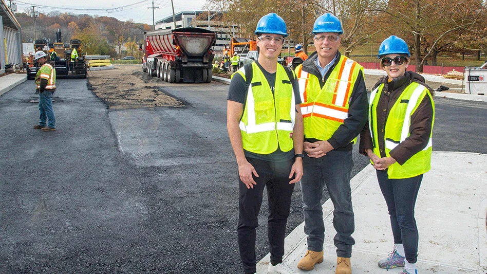 two men and one woman in yellow safety vests and blue helmets smile at asphalt paving site