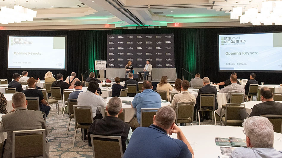 men and women seated at round tables in front of a diaz with two speakers seated on it